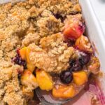 A white baking dish with a fruit crisp inside with peaches, blueberries, and a crisp topping. With a spoon inside the dish.