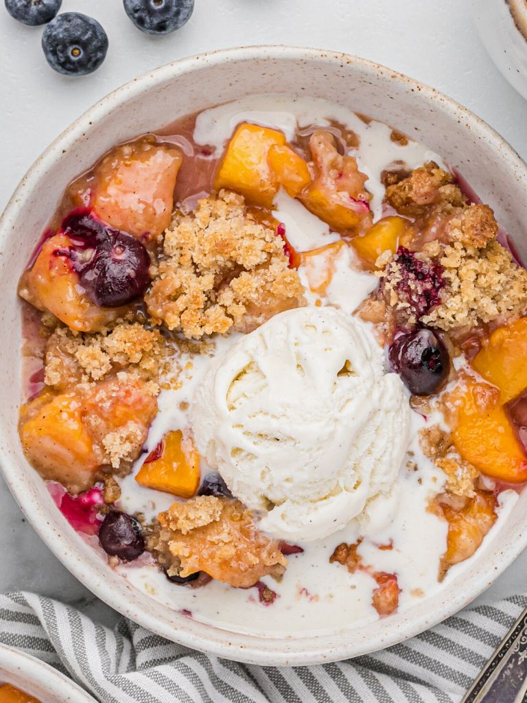 A bowl of fruit crisp with peaches and blueberries and topped with melted vanilla ice cream.