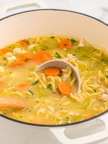 A white pot of soup with noodles, chicken, and carrots with a soup ladle inside the pot.