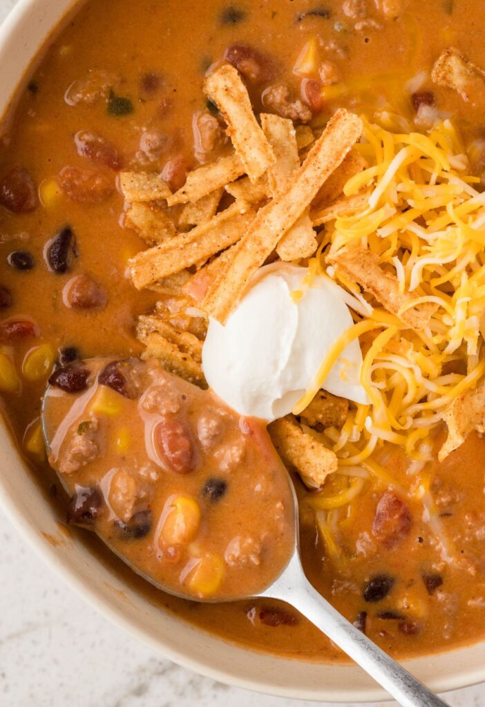 A bowl of taco soup with a spoon inside of it and with toppings. 