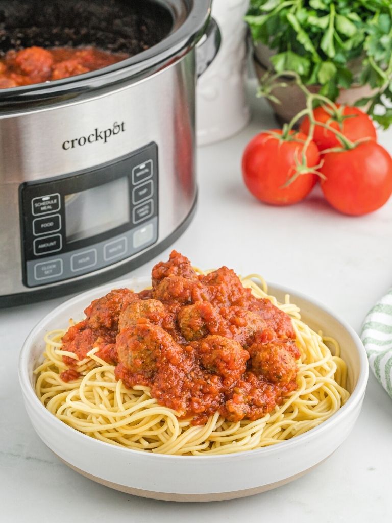 Slow cooker with a bowl of meatballs and spaghetti sauce in a white bowl in front of it. 