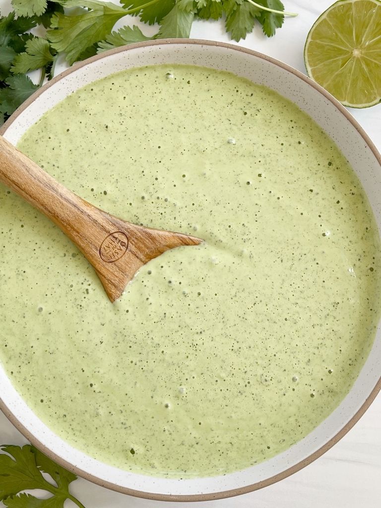 A bowl of green dressing with a wooden spoon inside of it. Lime pieces and cilantro pieces off to the side.