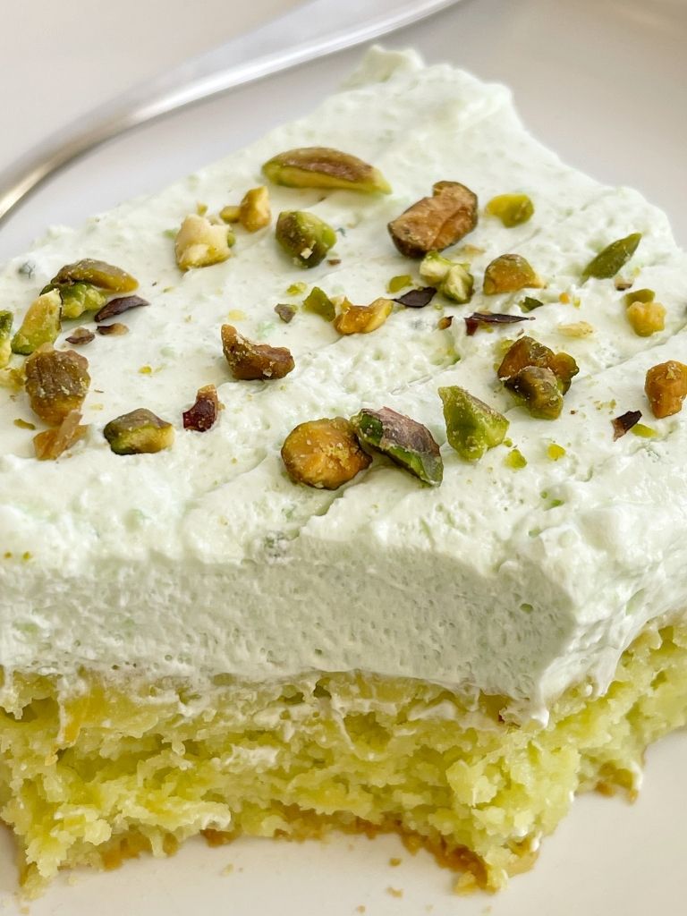 Slice of pistachio cake with frosting and pistachios on top of the cake slice.