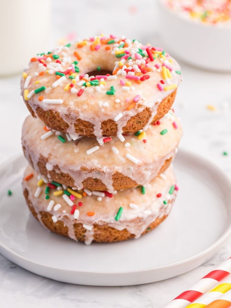 A stack of donuts with sprinkles on a white plate with color straws in the background.