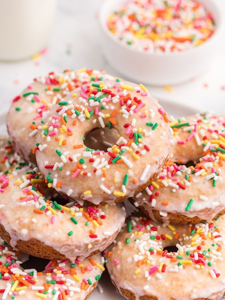 Funfetti Donuts stacked on a white plate with a bowl of sprinkles in the background.