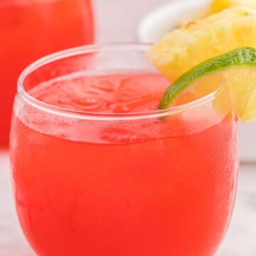 A glass of party punch in ice cubes and garnished with lime and pineapple slices on top of the glass.