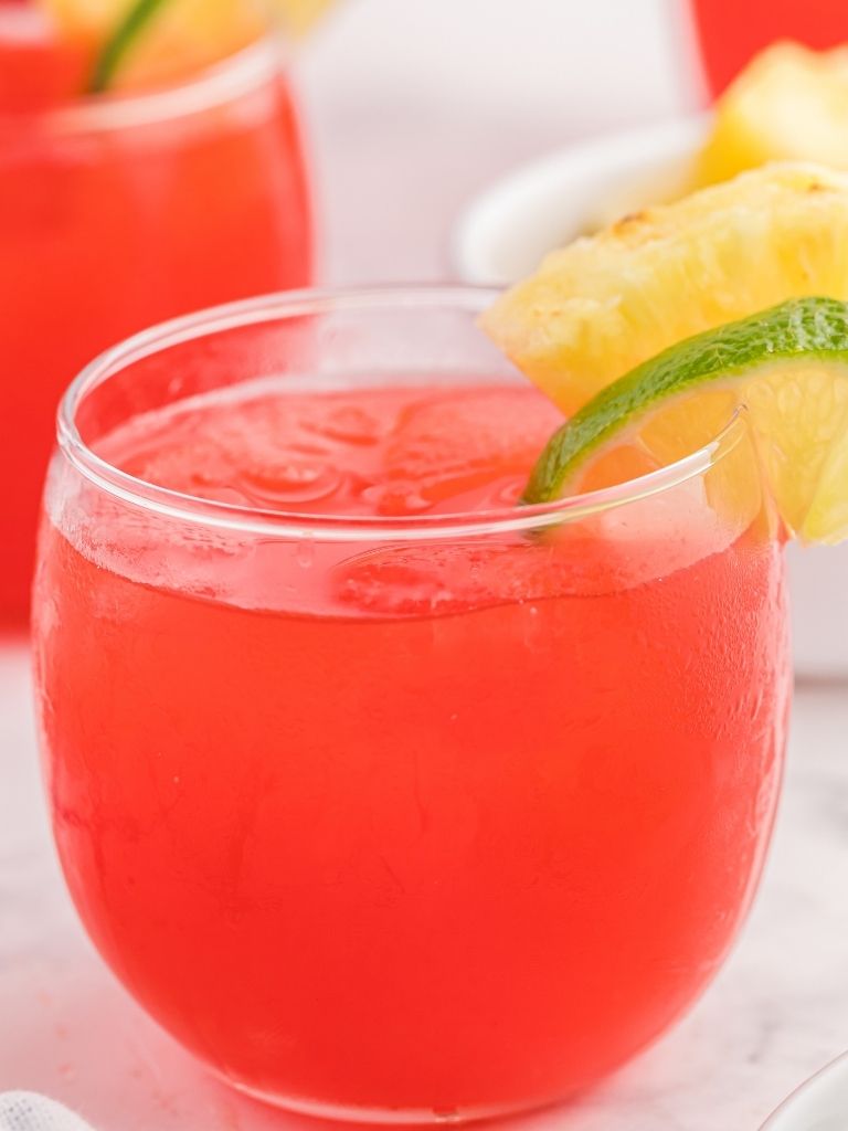 A glass of party punch in ice cubes and garnished with lime and pineapple slices on top of the glass. 