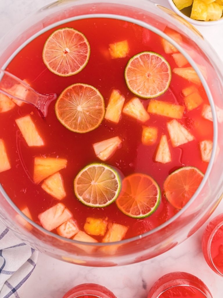 Punch bowl with red punch inside and garnished with lime slices and pineapple chunks on top.