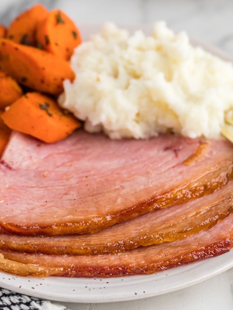 Ham slices on a white plate with carrots and mashed potatoes in the background. 