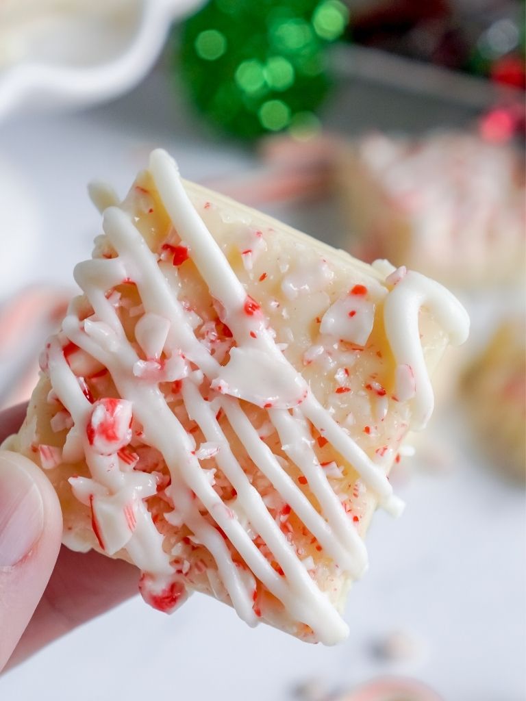 A hand holding one piece of fudge sprinkled with chocolate and crushed candy cane. Christmas lights in the background.