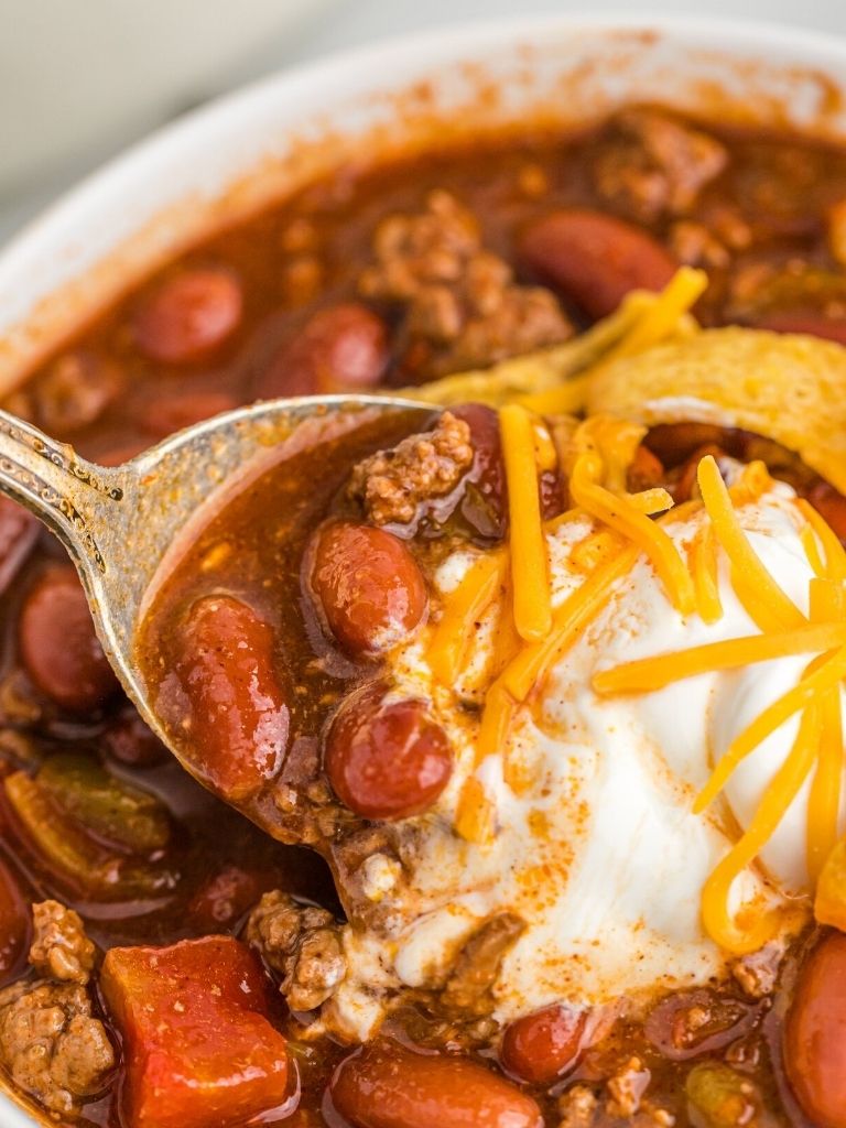 Chili inside a bowl topped with sour cream and cheeses. A spoon taking a spoonful of chili. 