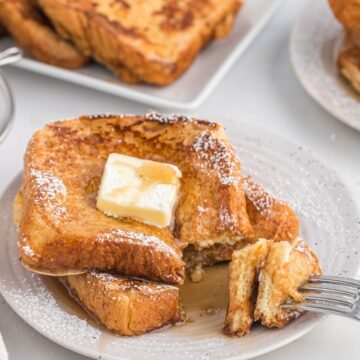 A plate of French toast with butter and a fork taking a bite from the stack.
