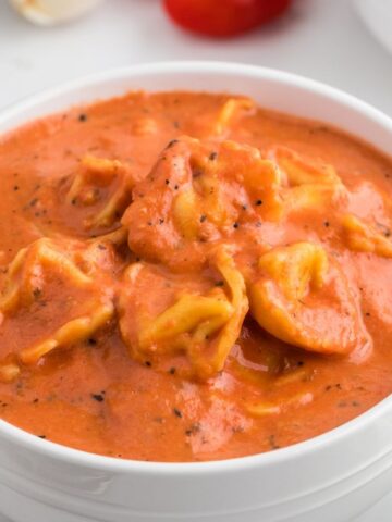 A white bowl with tomato tortellini soup inside of it on a white background.