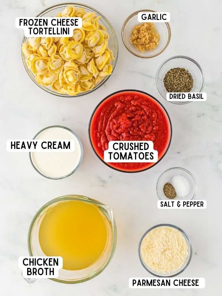 Ingredients laid on a white background for making creamy tomato soup. All ingredients are labeled in text. 