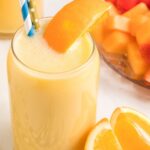 A glass with orange peach smoothie inside and a straw. Fruit in the background of the picture.