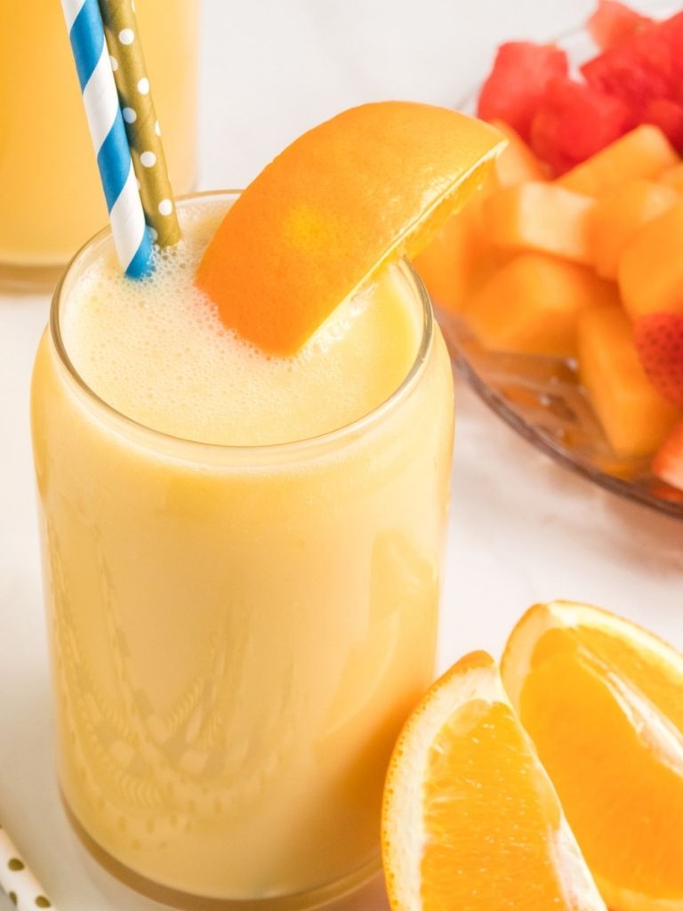 A glass with orange peach smoothie inside and a straw. Fruit in the background of the picture. 