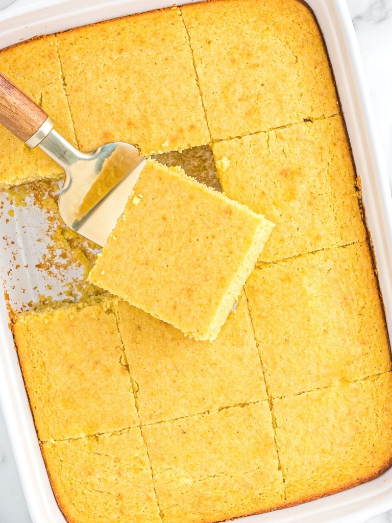 A white pan with cornbread inside of it with a spatula picking up a piece of it. 