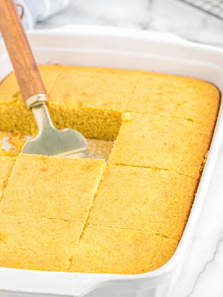White baking dish with cornbread inside of it and a serving spatula.