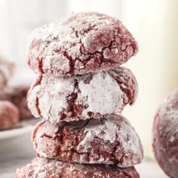 Stack of red velvet cookies coated in powdered sugar stacked with a milk glass behind them and a plate of cookies.