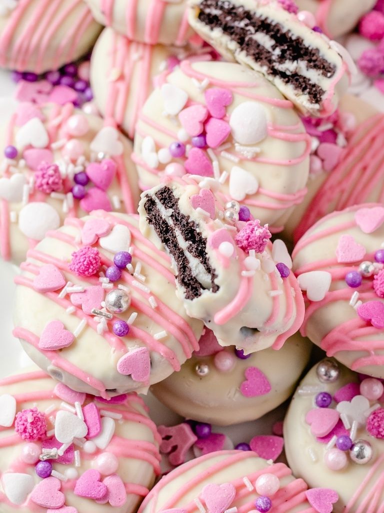 A pile of Oreo cookies covered in white chocolate and decorated with pink frosting and sprinkles. One Oreo is cut in half on the top of the pile to show the inside. 