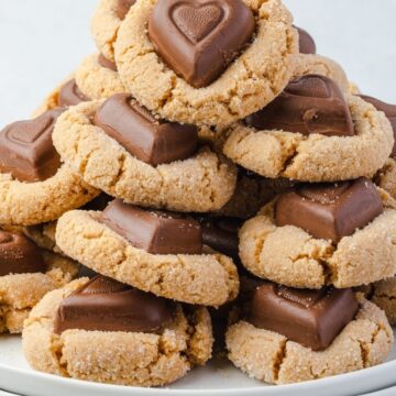 Stack of peanut butter cookies on a stack of three white plates. Each cookies is topped with a heart shaped chocolate on top.