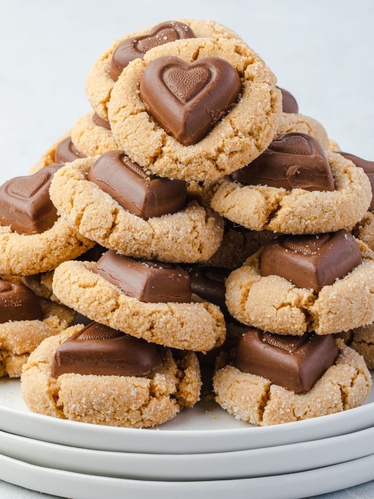 Stack of peanut butter cookies on a stack of three white plates. Each cookies is topped with a heart shaped chocolate on top. 