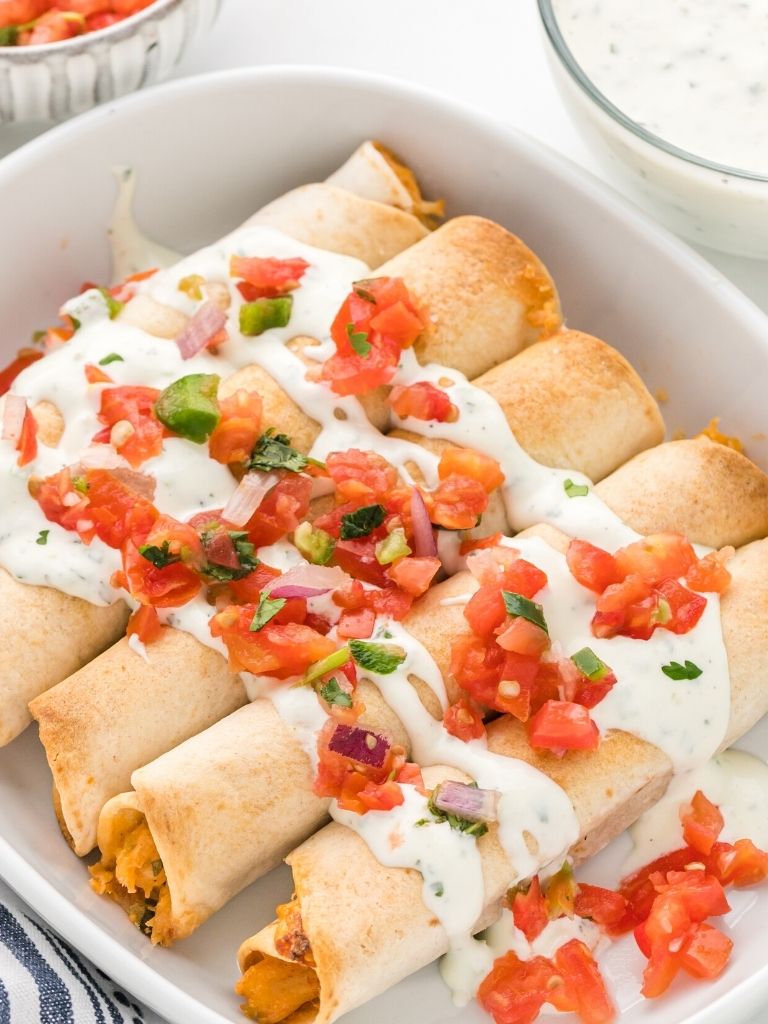 A white dish with taquitos inside of it and topped with sour cream and tomatoes. 
