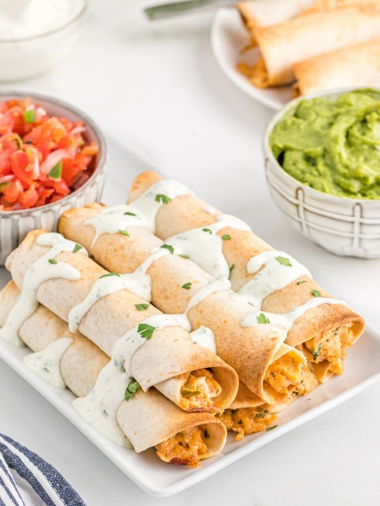 A picture of taquitos on a white plate drizzled with sauce. Blue towel and guacamole in the background.