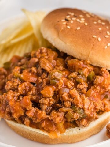 A white plate with a hamburger bun on it and chips in the background. Sloppy Joe meat on the bun.