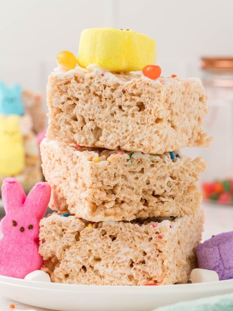 A stack of easter Krispie treats on a white plate with peeps bunnies at the side.