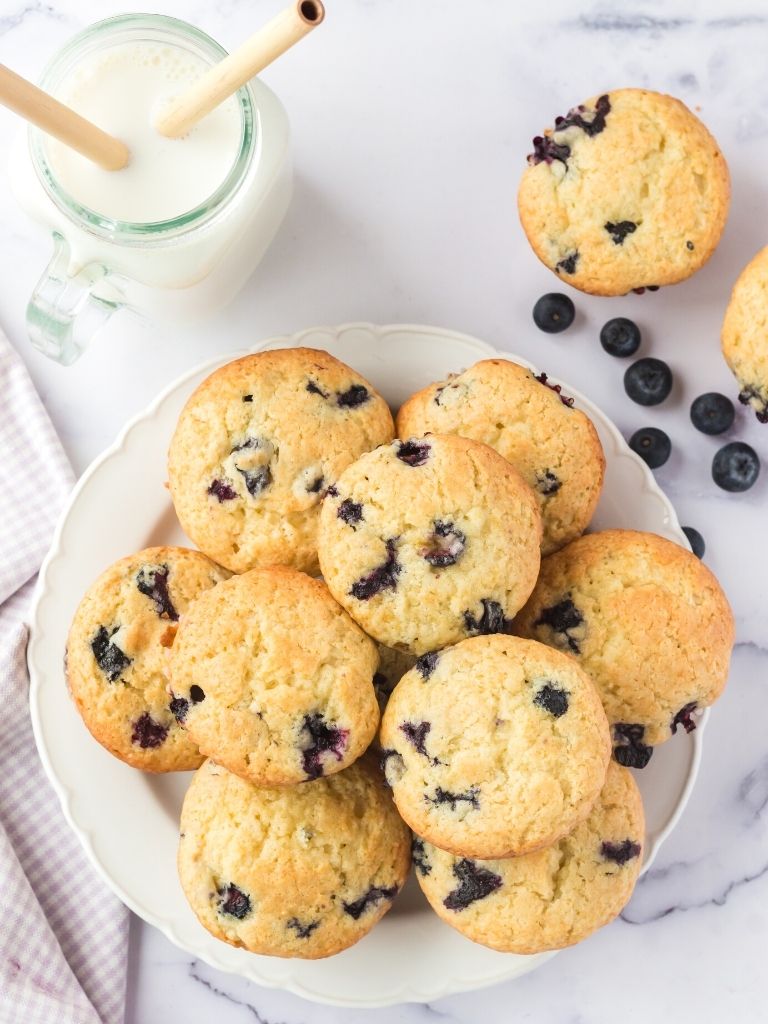 Overhead shot of muffins on a white plate with milk and blueberries in the background of the picture. 