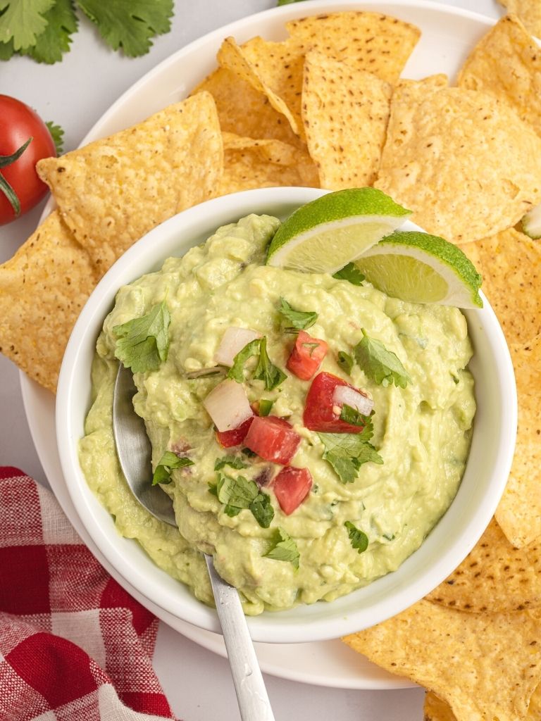 Overhead shot of a white bowl with guacamole in it, a spoon, and garnished with lime wedges and tomatoes. 