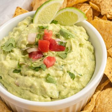 Creamy guacamole dip in a white bowl topped with tomatoes, onion, and lime wedges.