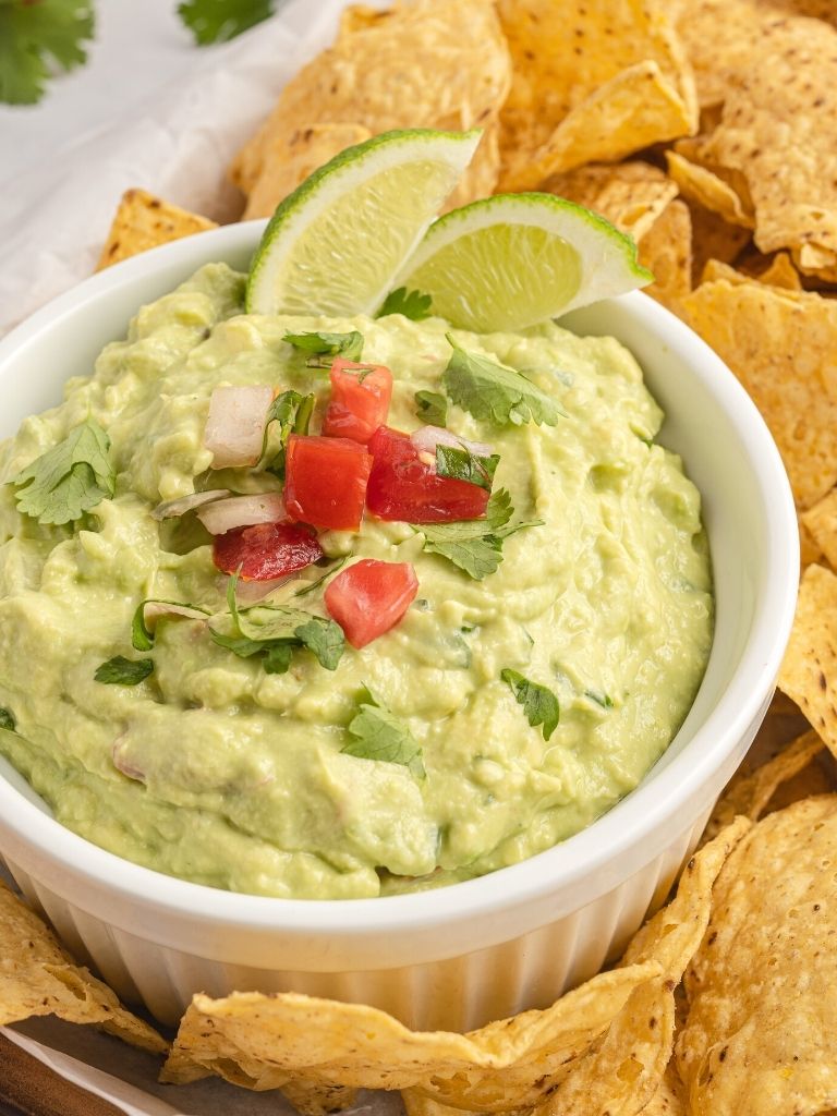 Creamy guacamole dip in a white bowl topped with tomatoes, onion, and lime wedges. 