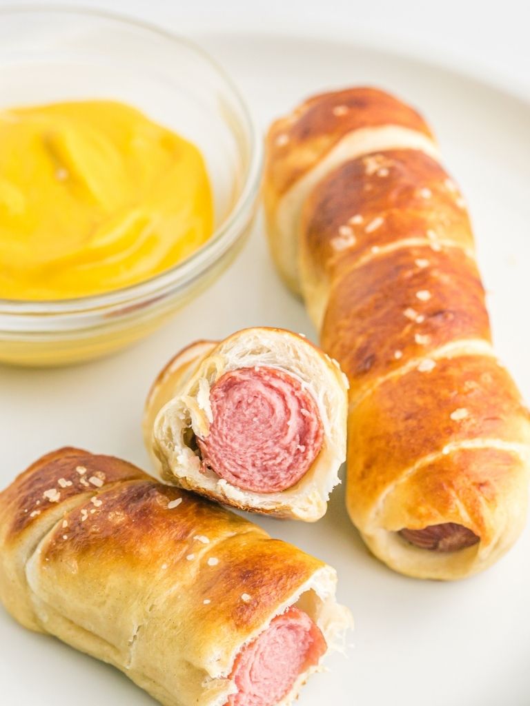 White platter with mustard dip and pretzel coated hot dogs with one cut in half to show the middle. 