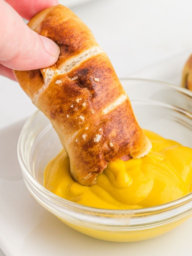 Mustard in a bowl with a pretzel hot dog being dipped in it. 