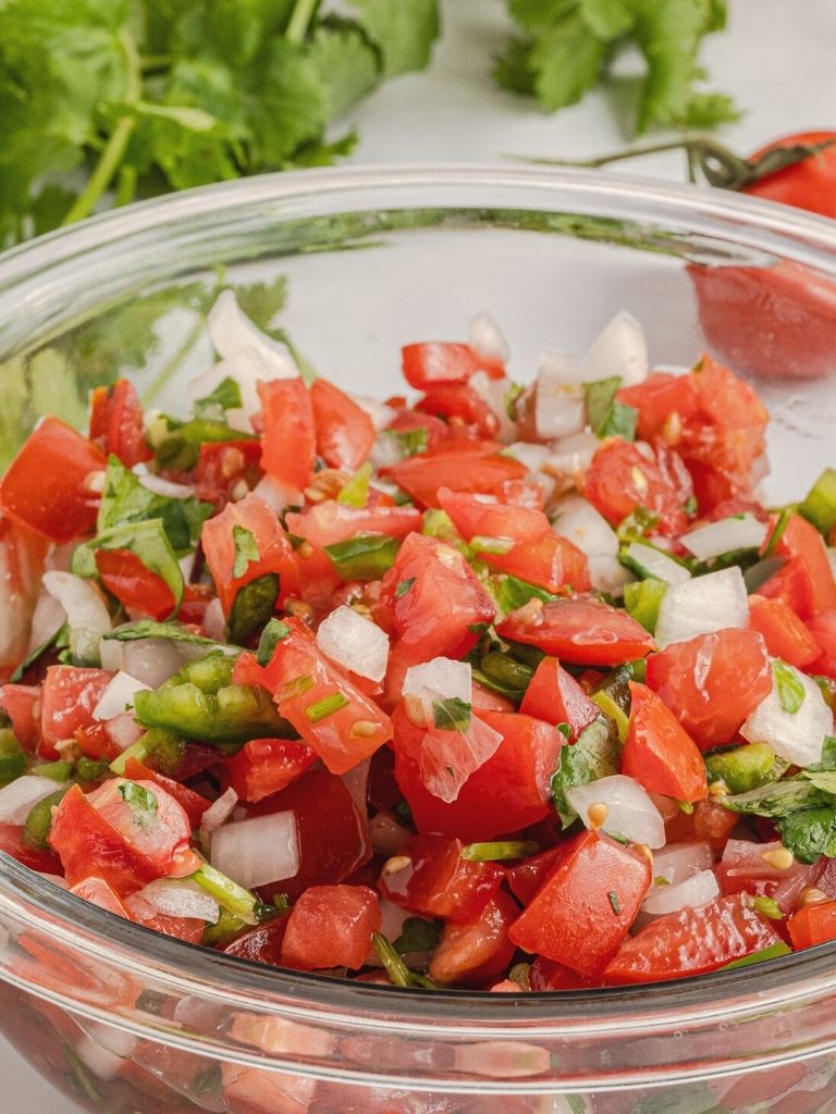 Tomato chopped salsa inside a clear glass bowl with cilantro in the background.