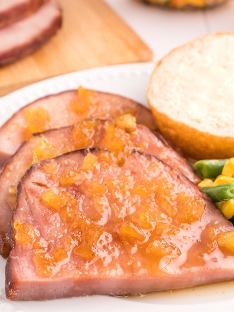 ham slices on a white plate with veggies and a roll. 