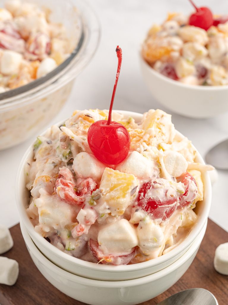 A serving of fluffy and creamy fruit salad inside a white bowl with a cherry on top.
