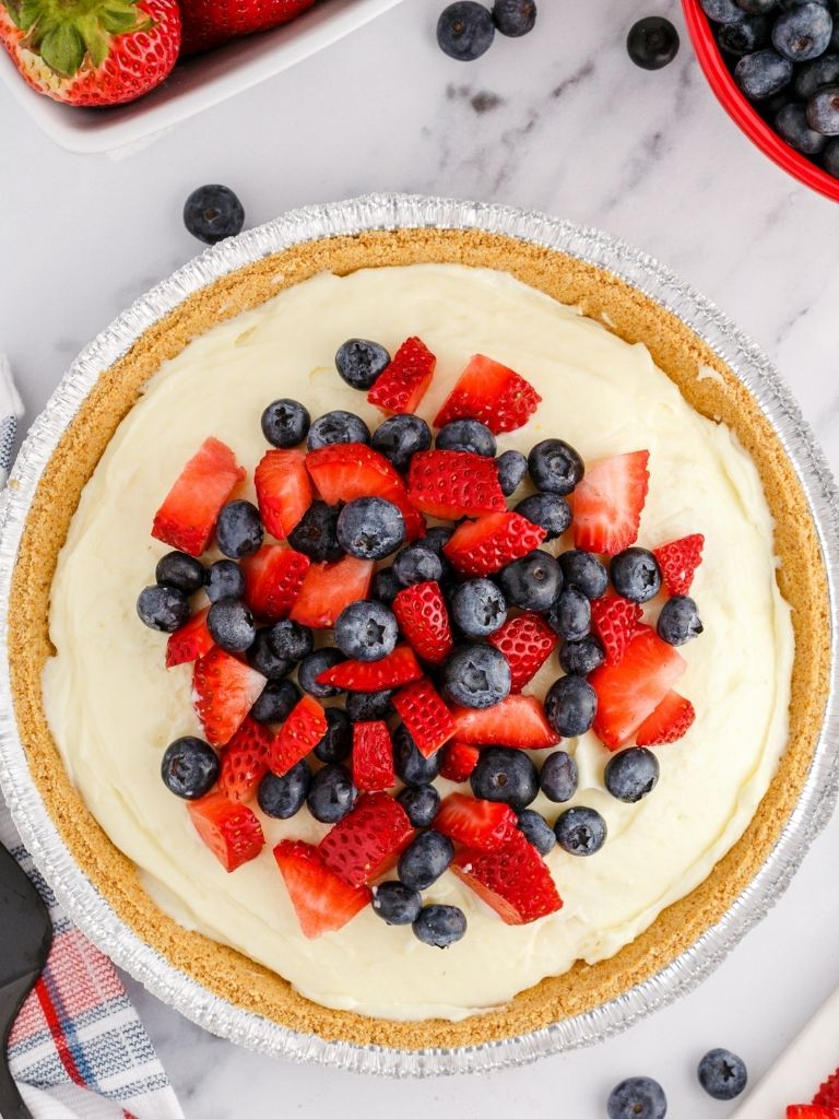 A berries cream pie topped with chopped blueberries and strawberries with fresh berries in the background of the photo. 
