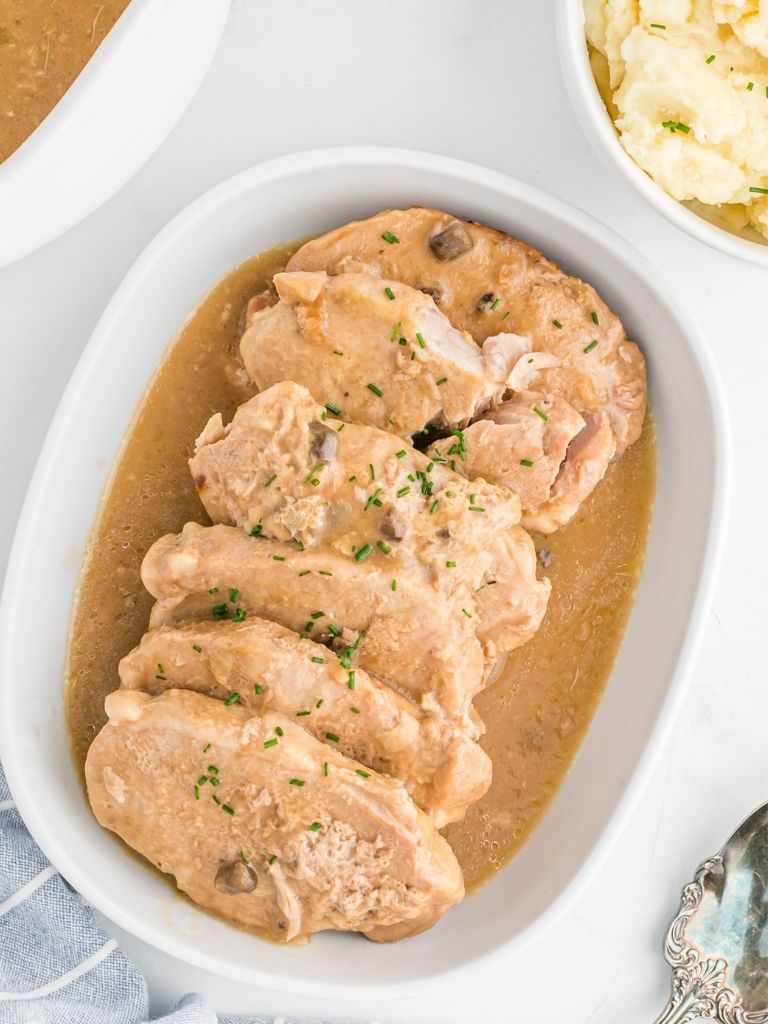 Mushroom pork chops inside a white serving dish topped with chopped chives and gravy inside a bowl next to it. 