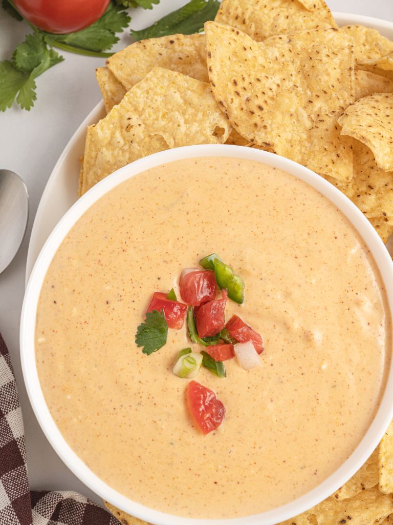 A white bowl with queso in it and topped with tomatoes with chips and cilantro in the background.