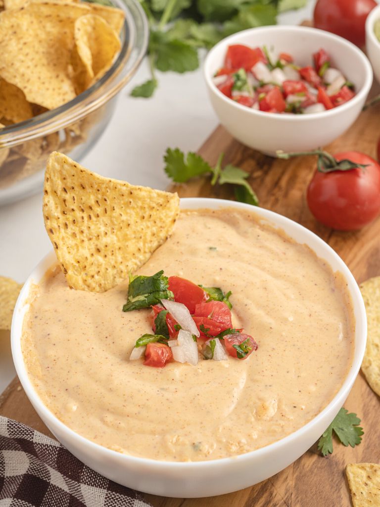 Queso with a chip dipped in it and chips, pico de Gallo in the background of the photo.