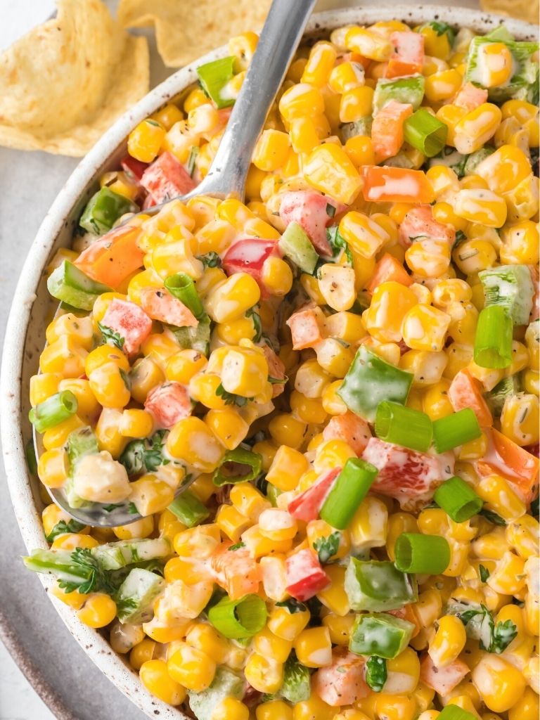White bowl with corn salad inside of it with bell peppers and a silver spoon.