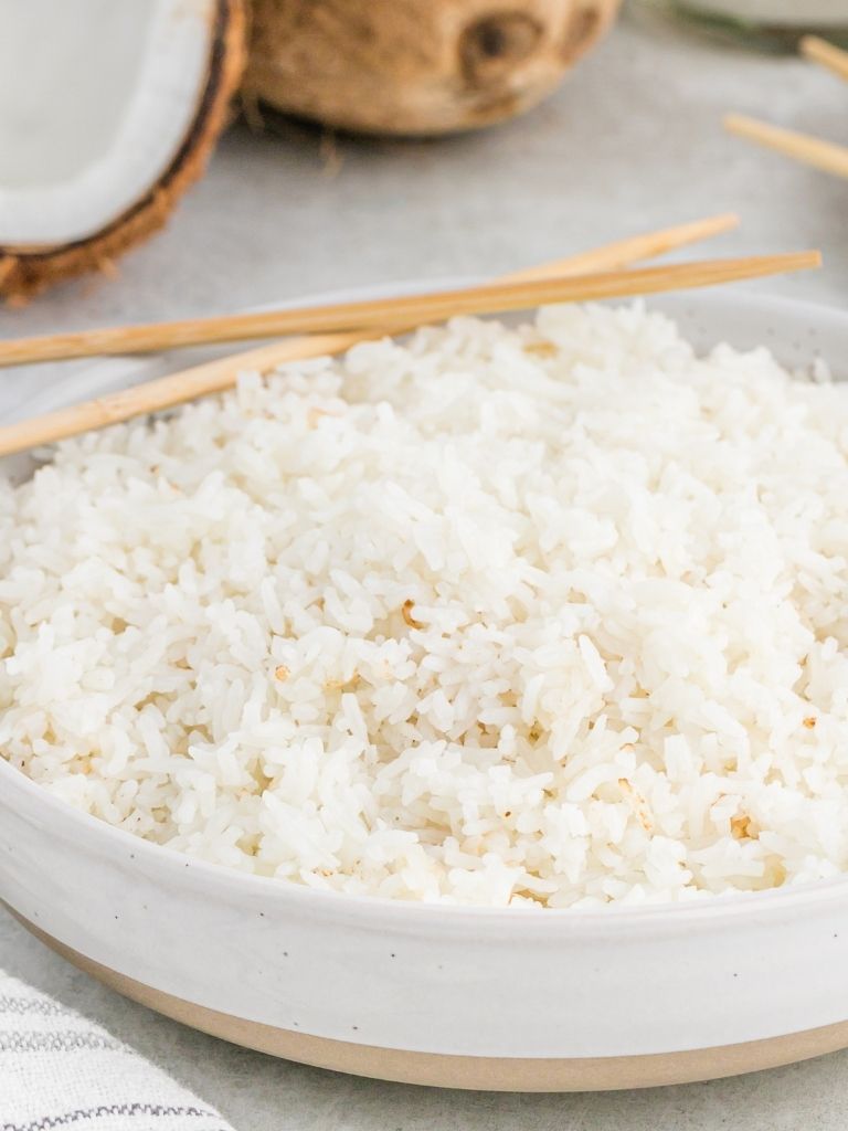 A white bowl of coconut rice in rice cooker with a coconut in the background of the picture.