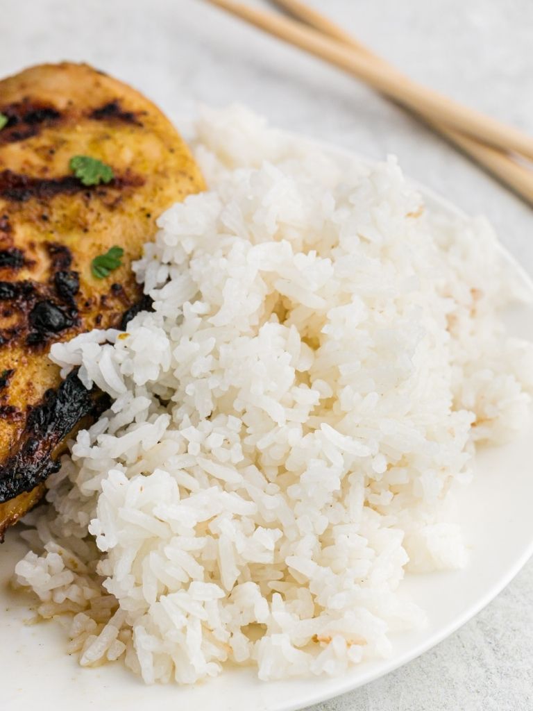 A serving of coconut rice on a white plate with grilled chicken in the background