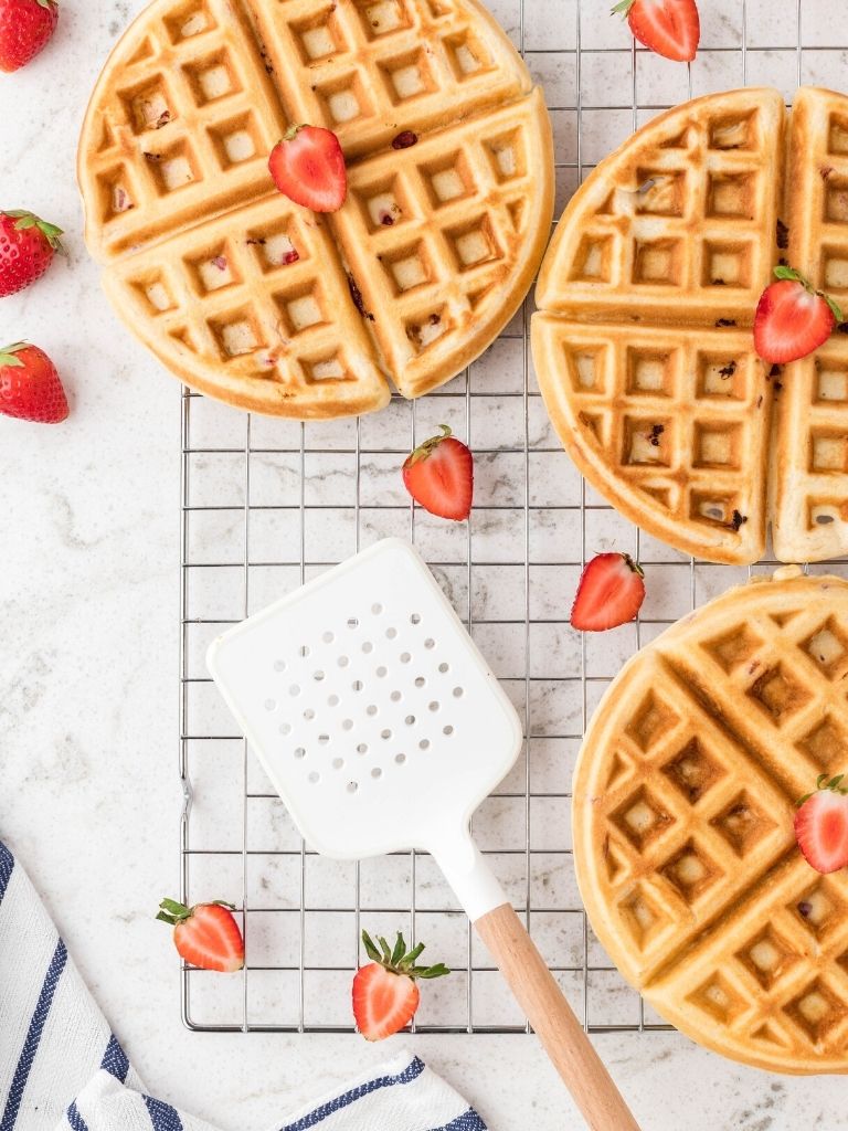 Cooling rack of waffles topped with strawberries with a white spatula next to it. 