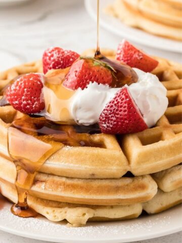 Stack of strawberry waffles on a white plate with whipped cream and maple syrup being drizzled over the top of the stack.