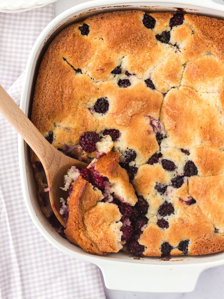 A white baking dish with blackberry cobbler and a wooden spoon taking a scoop of cobbler inside the baking dish. 