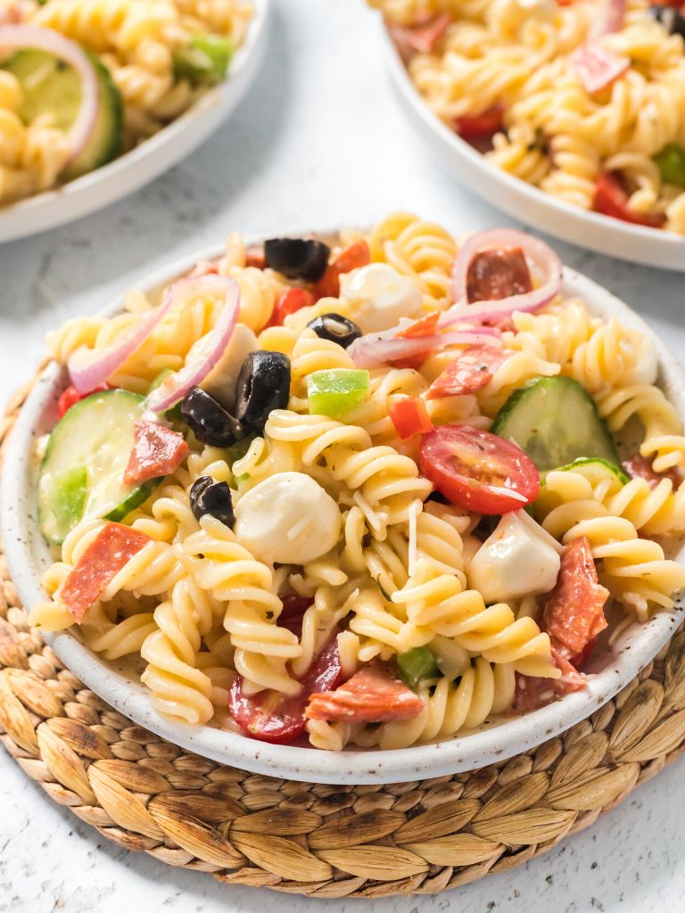 Pasta salad in a white bowl on top of a brown wooded placemat. 